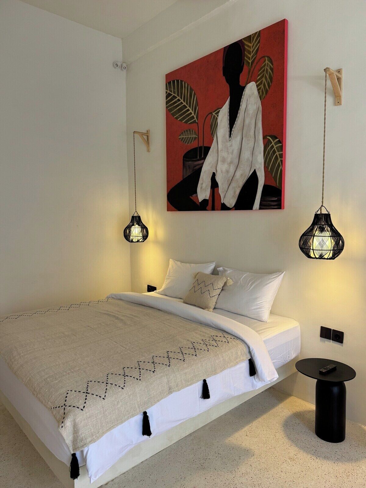 A bedroom with a bed, a painting, two pendant lights, and a small table.