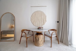 Quarto com mesa redonda, duas cadeiras, um detalhe de macramê pendurado na parede, espelho e janela com cortinas.