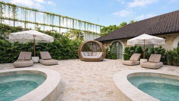 A pool area with lounge chairs and umbrellas, surrounded by greenery and a building with a tiled roof.