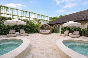 Área de piscina com espreguiçadeiras e guarda-sóis, cercada por vegetação e um edifício com telhado em azulejos.