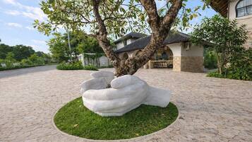 A tree with a unique sculpture of a hand in a planter, a paved driveway, and a building with a tiled roof in the background.