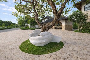 Uma árvore com uma escultura singular de uma mão em um vaso, um caminho de paralelepípedos e um edifício com telhado cerâmico ao fundo.