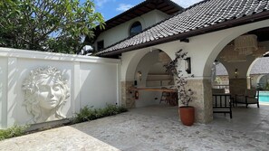 Área externa coberta com piso de pedra, parede branca com escultura em relevo e espaço de estar com cadeiras e mesa.