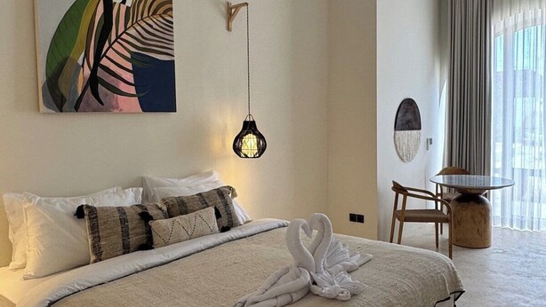 A hotel room with a bed, a painting, a hanging light fixture, a dining area with a table and chairs, and a view of the outdoors through a window. - Brand New Apartments in Sanur+500m to Ocean+Pool (Kota Denpasar)