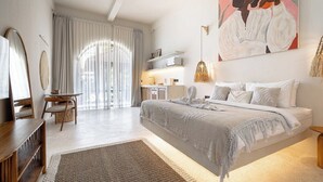 Ein Schlafzimmer mit einem großen Bett, einem Schreibtisch und einem Sessel. An der Wand hängt ein Gemälde, und ein Fenster mit Vorhängen ist zu sehen.