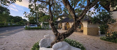 A tree with a unique sculpture of hands embracing it, set in a landscaped area with a cobblestone path and a building in the background.