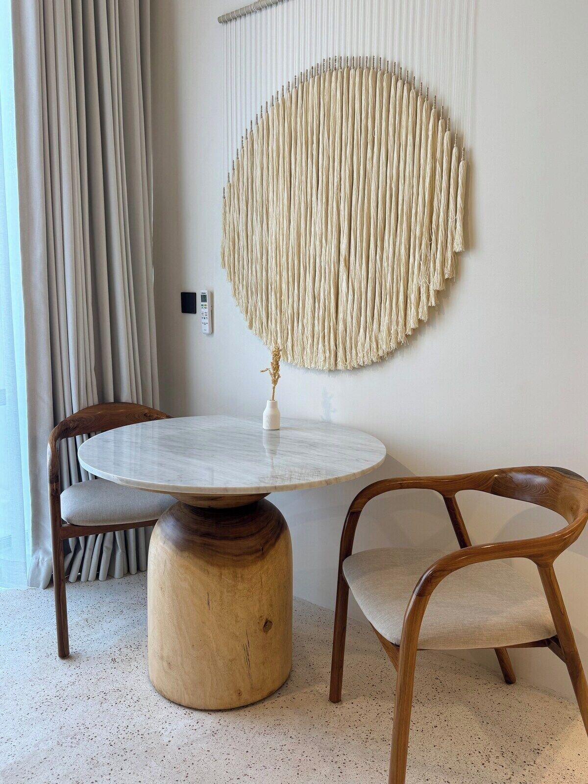 Een kamer met een ronde houten tafel, twee stoelen, een macramé wanddecoratie en een gordijn.