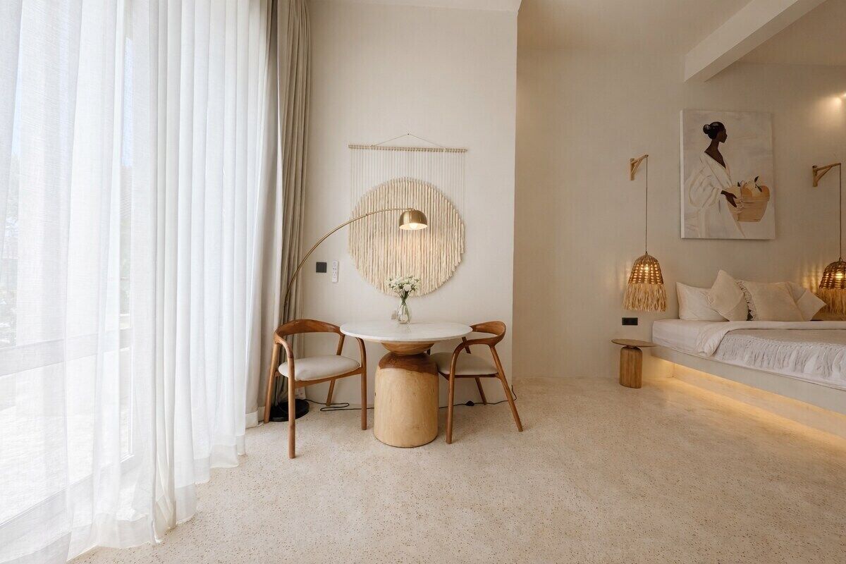 A modern bedroom with a round table, two chairs, a bed with white bedding, a wall-mounted lamp, and a framed artwork.