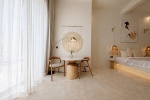 A modern bedroom with a round table, two chairs, a bed with white bedding, a wall-mounted lamp, and a framed artwork. - New Cozy 1BR Next to the Ocean w Pool+Patio+Garden (Kota Denpasar)
