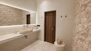 A modern bathroom with a stone-textured wall, a wooden door, a large mirror, a freestanding sink, and a toilet. - New Cozy 1BR Next to the Ocean w Pool+Patio+Garden (Kota Denpasar)