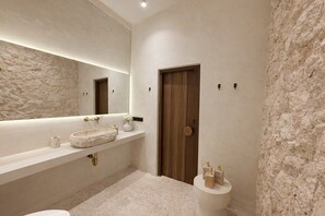 A modern bathroom with a stone-textured wall, a wooden door, a large mirror, a freestanding sink, and a toilet. - New Cozy 1BR Next to the Ocean w Pool+Patio+Garden (Kota Denpasar)
