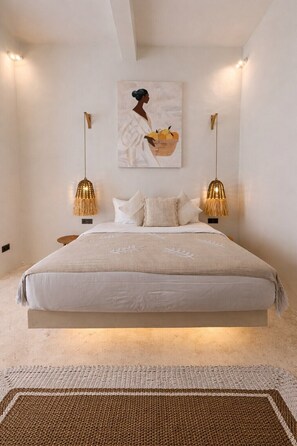 A bedroom with a bed, two wall-mounted lamps, a painting, and a textured rug. - New Cozy 1BR Next to the Ocean w Pool+Patio+Garden (Kota Denpasar)
