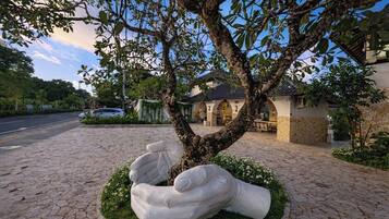 Un albero con una scultura particolare di mani che lo avvolgono, inserito in un’area verde con un sentiero in pietra e un edificio sullo sfondo.
