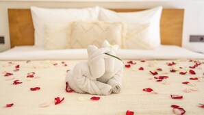 Una cama impecablemente tendida con un cabecero blanco, una toalla enrollada con forma de conejo y pétalos de rosa esparcidos sobre el edredón.
