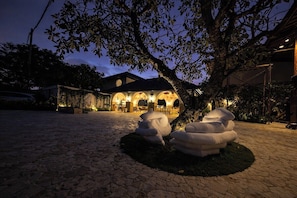A building with a circular driveway, a tree with a well-lit area underneath, and a structure with arches in the background. - Mezzanine Style Apartment in Boutique Hotel+Pool (Kota Denpasar)