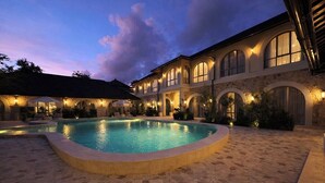 Una piscina rodeada por un patio de piedra, con un edificio que tiene ventanas de arco y luces exteriores.