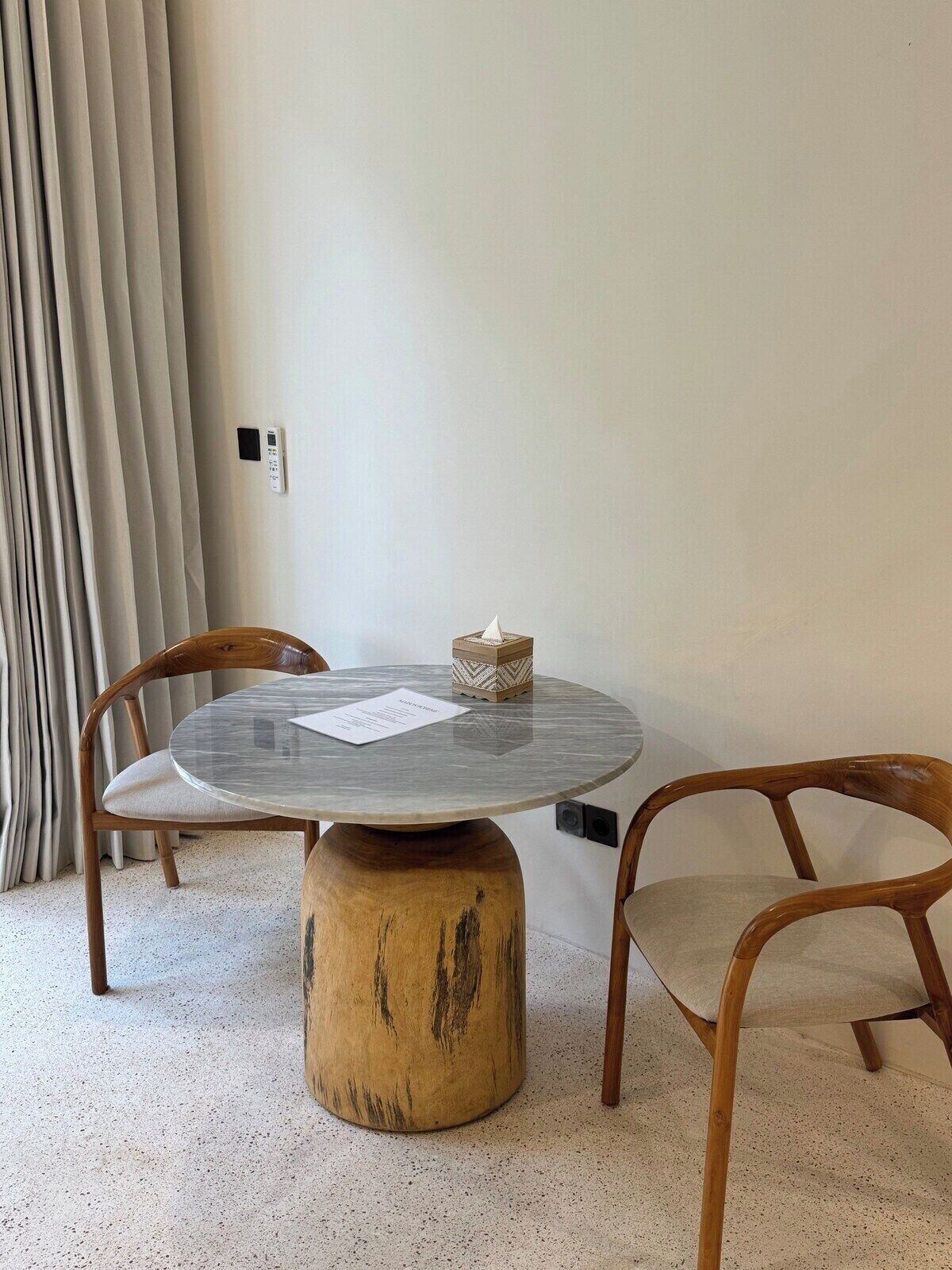 A round table with a wooden base, two wooden chairs, a small decorative item, and a piece of paper on the table.