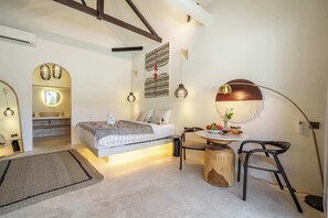 A modern bedroom with a bed, bedside table, and a desk with a chair. There is a mirror, a hanging light, and a wall-mounted artwork. - New Apartments in Sanur+5 min to Ocean+Pool (Kota Denpasar)