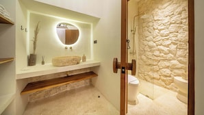 Un baño moderno con pared de piedra, estante de madera, espejo redondo e inodoro empotrado.
