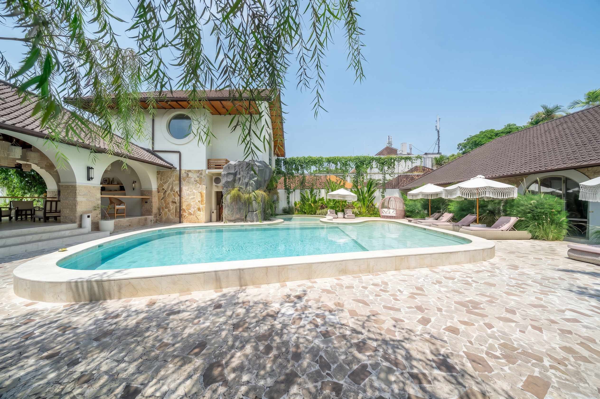 A swimming pool with a stone deck, surrounded by lounge chairs and umbrellas, with a building featuring arches and a tiled roof in the background.
