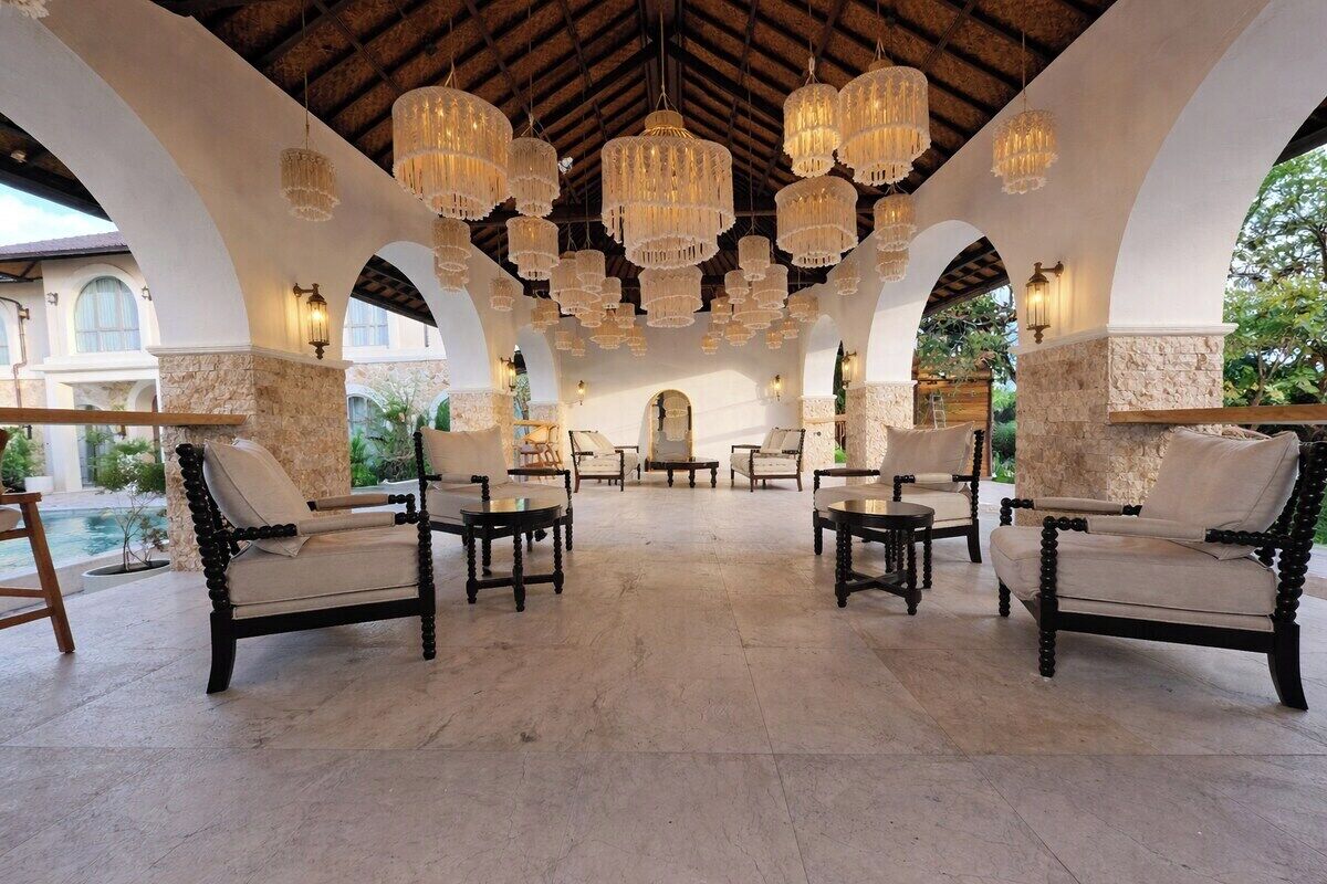Un área de estar al aire libre con múltiples sillas y mesas, suelo de piedra y candelabros colgantes.