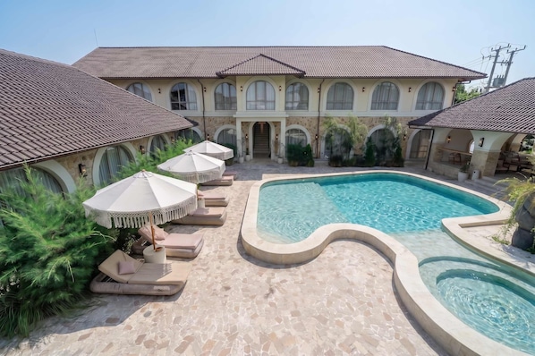 Área de piscina com espreguiçadeiras e guarda-sóis, e um edifício grande com janelas em arco ao fundo.