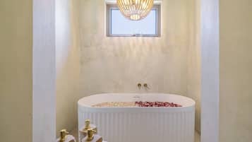 Un bagno moderno con una vasca freestanding, una finestra sopra la vasca e un lampadario decorativo.