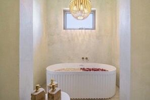 Un bagno moderno con una vasca freestanding, una finestra sopra la vasca e un lampadario decorativo.