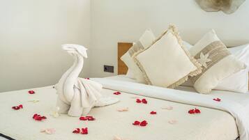 A hotel room with a bed adorned with a towel shaped like a swan, scattered with rose petals, and surrounded by white pillows with fringe details.