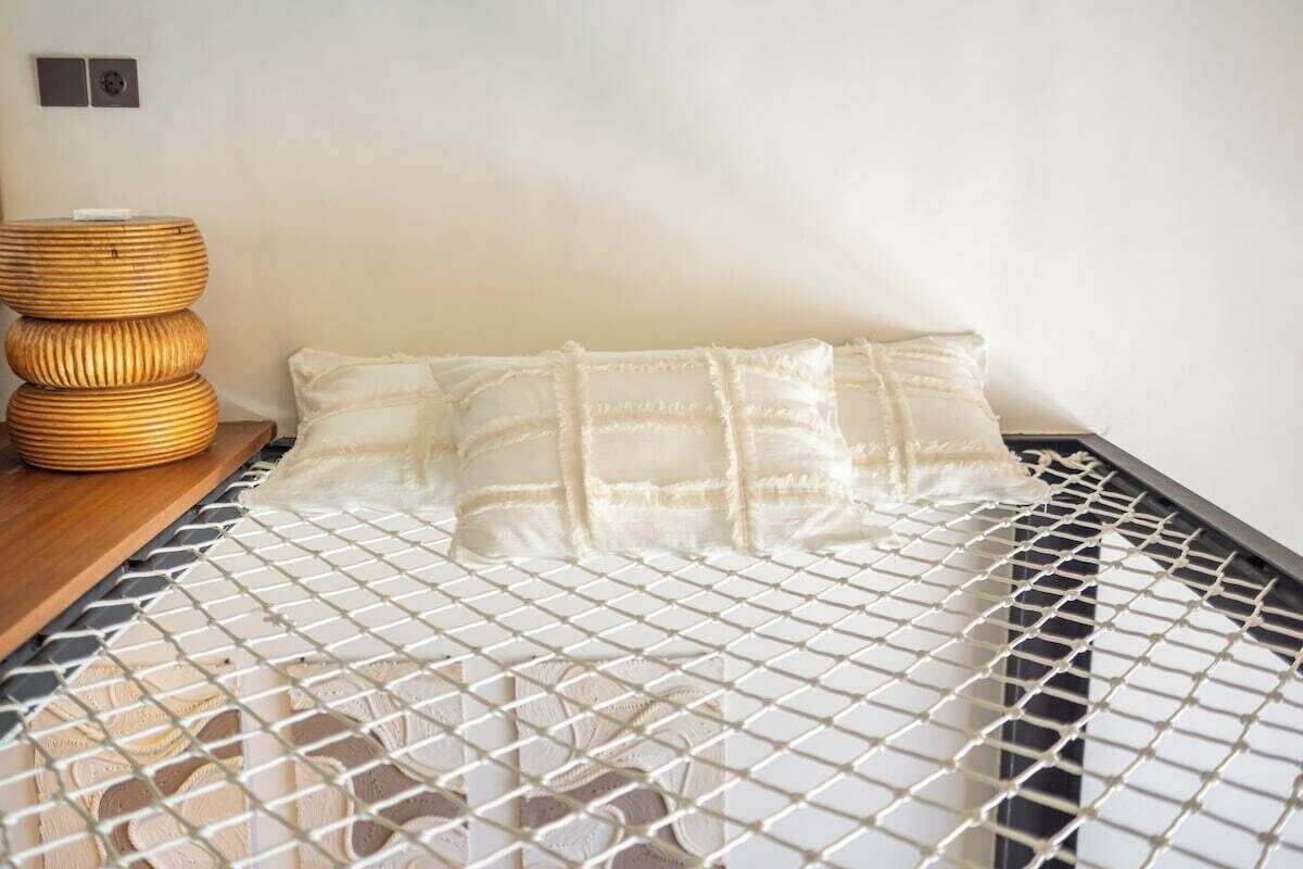 Een bed met een witte matras en kussen, een geweven nachtkastje en een wandlamp.
