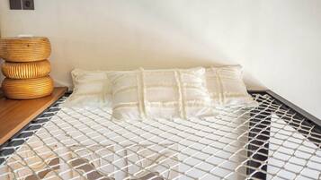 Een bed met een witte matras en kussen, een geweven nachtkastje en een wandlamp.