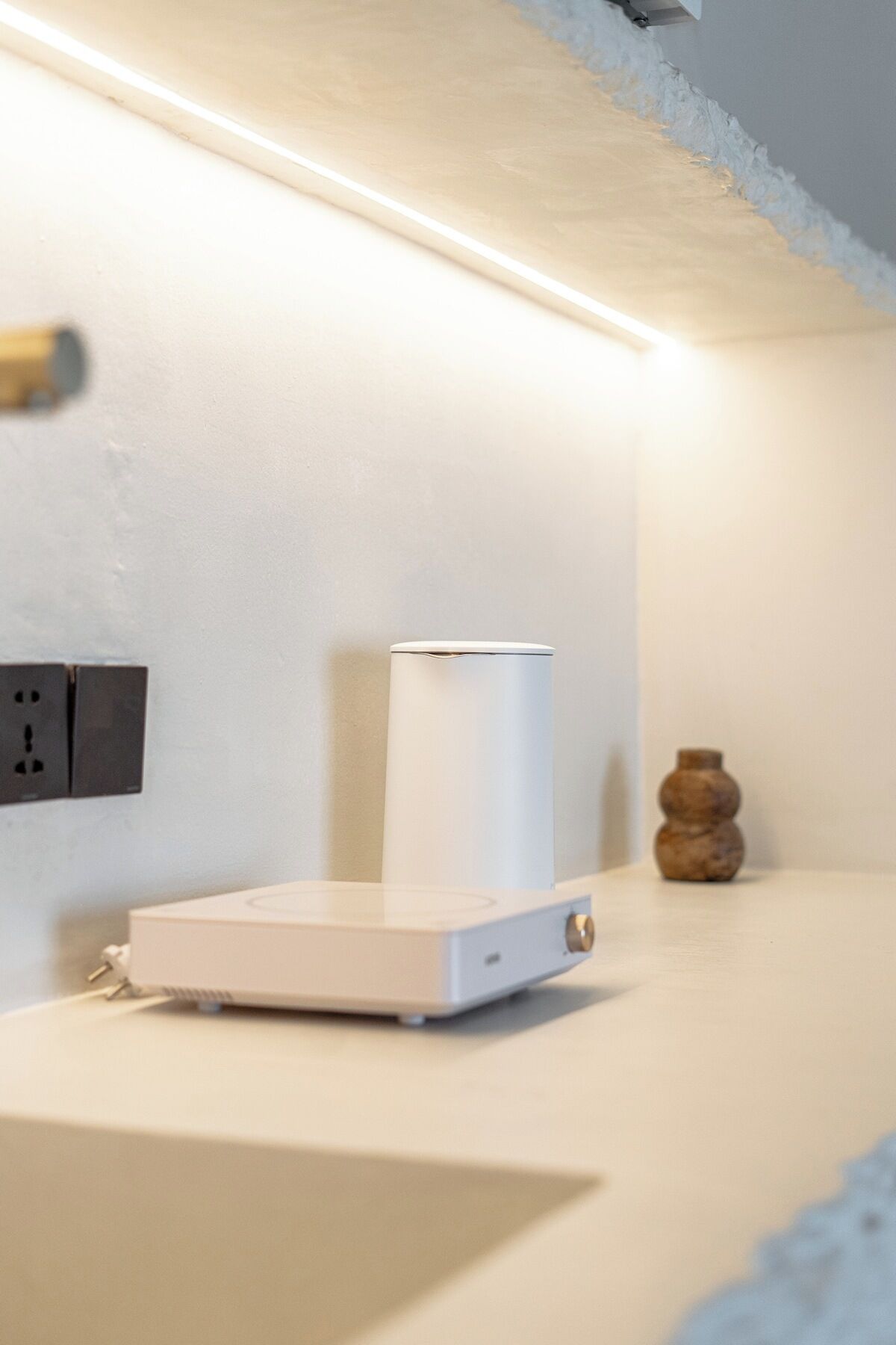 A white device on a countertop, a wall socket, and a small vase.