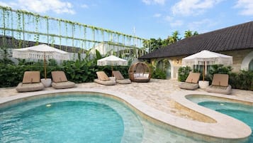 A pool area with lounge chairs and umbrellas, surrounded by greenery and a building with a tiled roof.