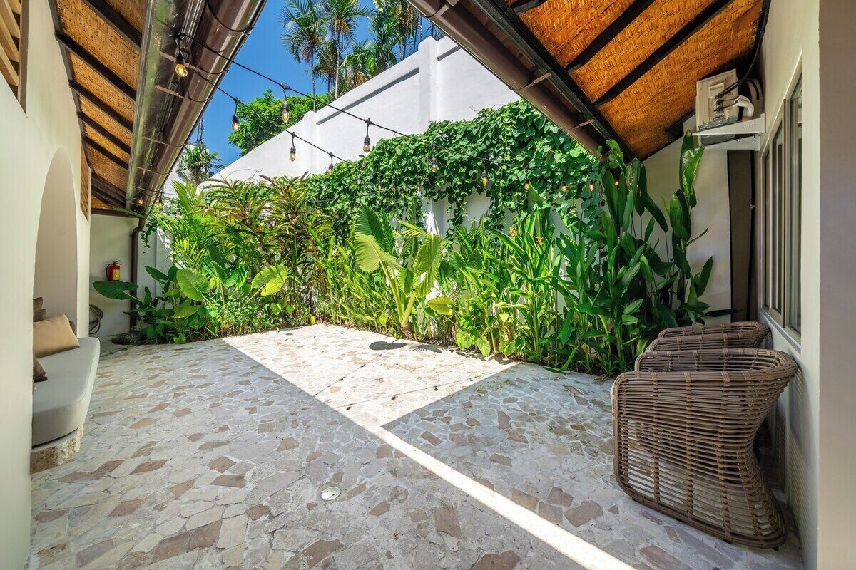 A covered outdoor area with a stone floor, a wicker chair, and lush greenery.