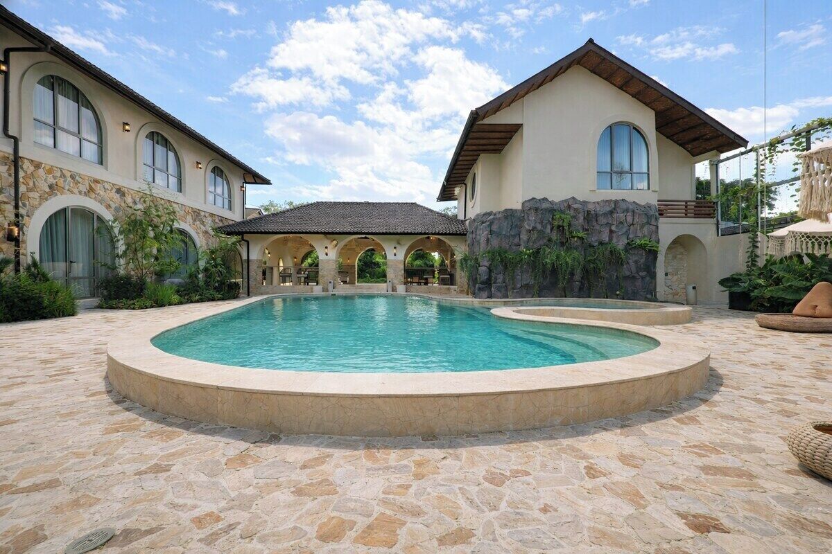 A pool area with a stone floor, a building with arched windows, and a covered patio.