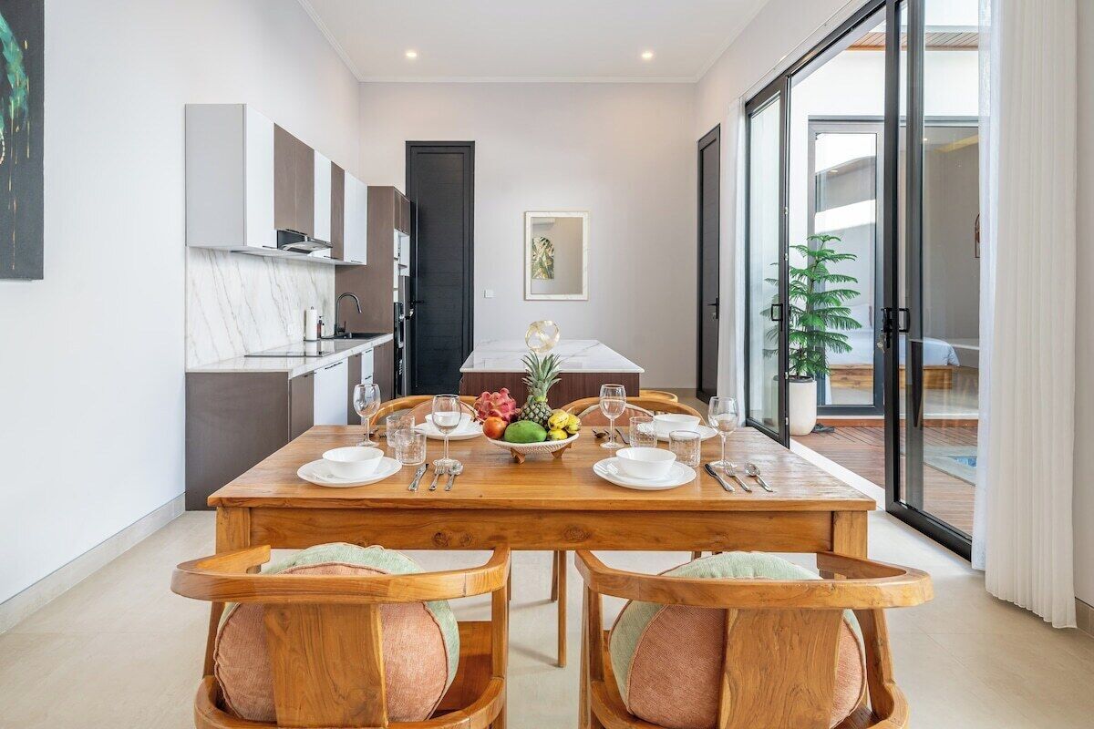 A dining area with a wooden table set for four, a kitchen in the background, and a sliding glass door leading to an outdoor area.