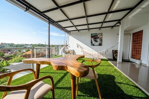 A balcony with a wooden table and chairs, a hammock, and a view of the city. - Chic 3BR in Nusa Dua w Ocean View+Cinema+Rooftop (Kabupaten Badung)