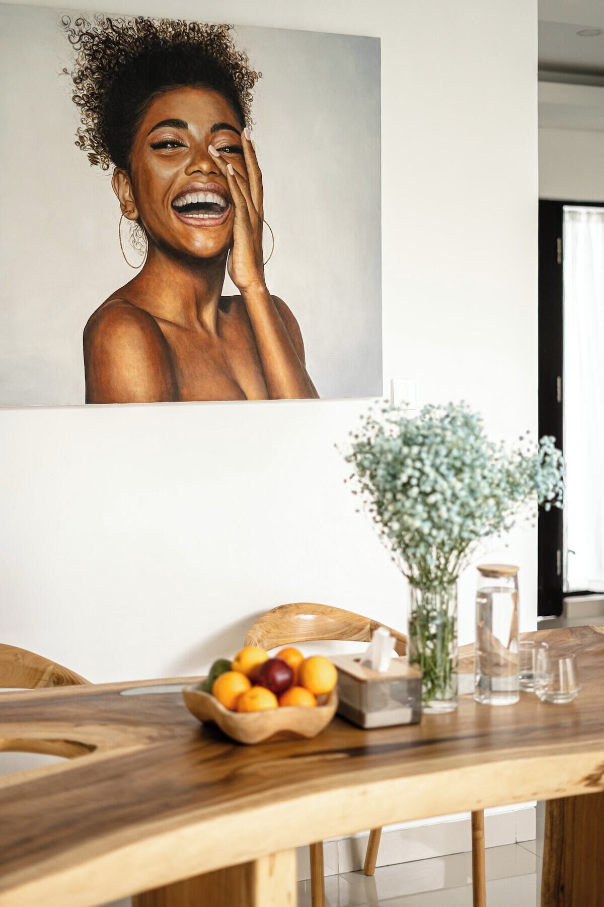 Un coin repas avec une table en bois, un saladier de fruits, un vase de fleurs et un tableau accroché au mur.