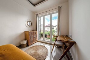 A room with a bed, a wooden table, a chair, a woven rug, a cabinet, and a large window with a view of houses and greenery. - Chic 3BR in Nusa Dua w Ocean View+Cinema+Rooftop (Kabupaten Badung)