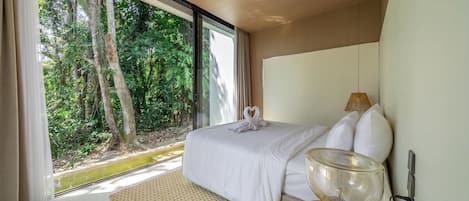 A bedroom with a large window, a bed with white bedding, a bedside table with a lamp, and a view of a forest.