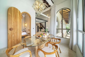 A modern dining area with a glass table surrounded by wooden chairs, a chandelier, and a view of a garden through large windows. - Cozy 2-Storey Flat by Sanur Beach w Pool & Office (Kota Denpasar)