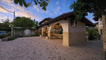 A stone-paved courtyard with a covered outdoor seating area, a stone wall with a carved face, and a clear sky.