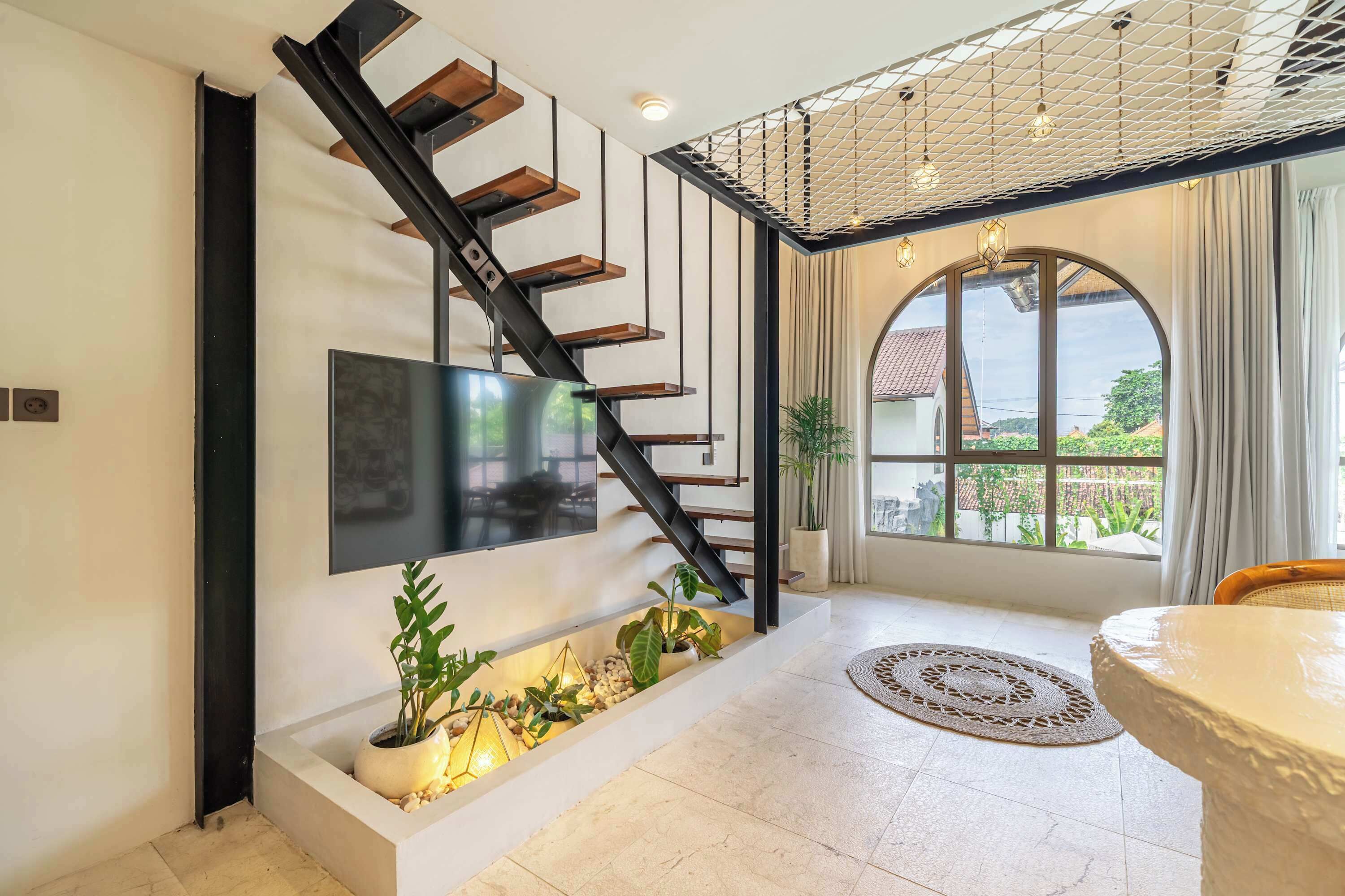 A modern interior with a staircase, a flat-screen TV, a dining area, and a view through a large window.