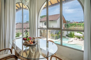 Zona pranzo con tavolo in vetro e sedie, una ciotola di frutta e pasticcini, e vista su una piscina e case attraverso ampie finestre.