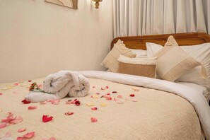 Un lit bien fait, avec une serviette roulée et des pétales de rose éparpillés, une tête de lit en bois et une table de chevet sur laquelle se trouve un panier.