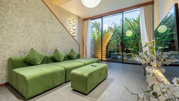 Un salon moderne avec un canapé d’angle, un petit pouf et une grande fenêtre donnant sur un paysage verdoyant.