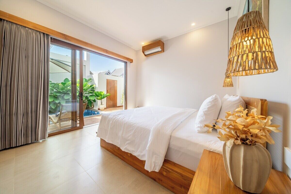 A bedroom with a bed, a wooden bedside table, a vase with flowers, a hanging light fixture, and a sliding glass door leading to a balcony with plants.