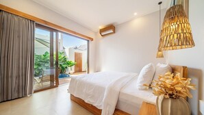 A bedroom with a bed, a wooden bedside table, a vase with flowers, a hanging light fixture, and a sliding glass door leading to a balcony with plants. - Designer 3BR Villa by Sanur Beach • Pool & Rooftop (Denpasar Selatan)