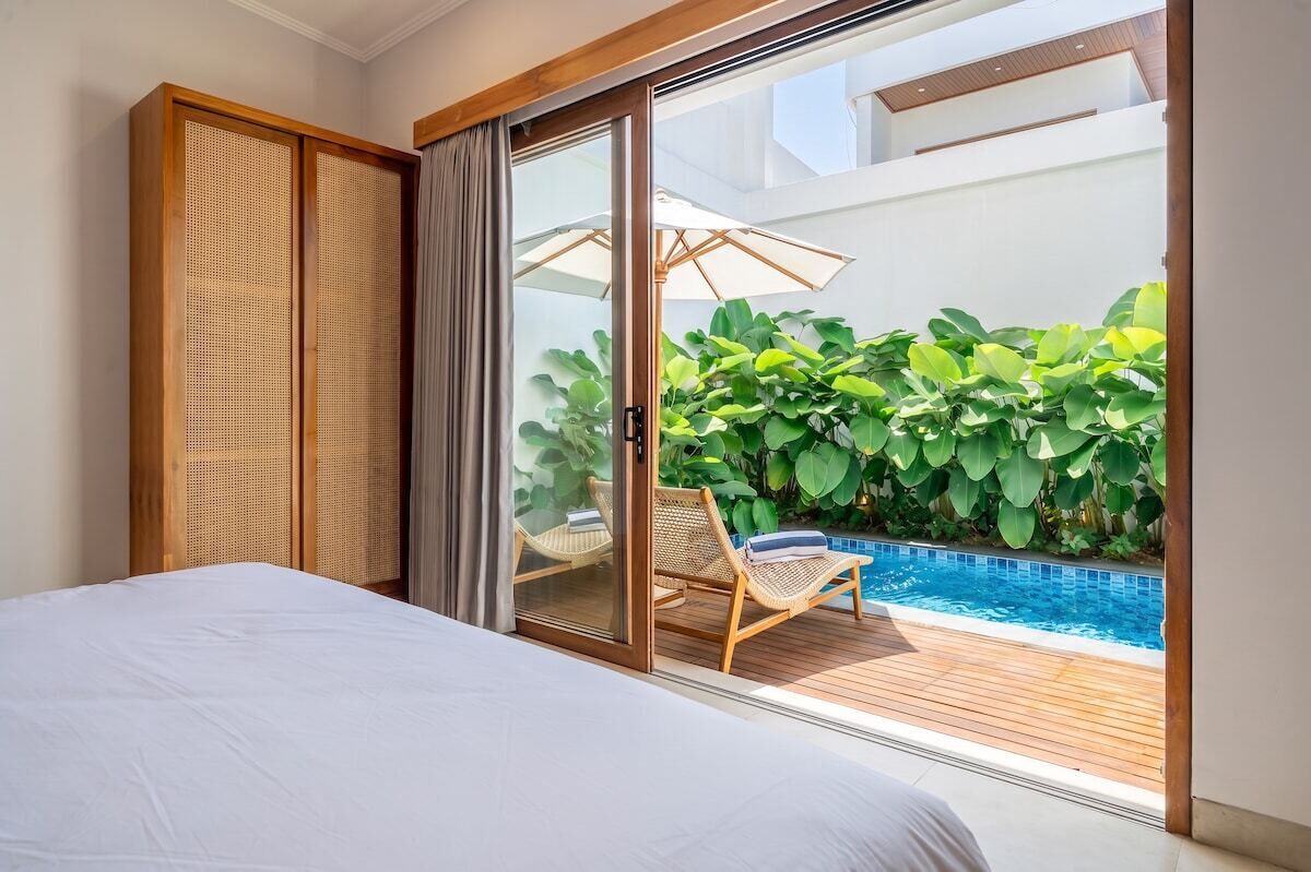 A bedroom with a large bed, sliding glass doors leading to a balcony with a pool and lush greenery.