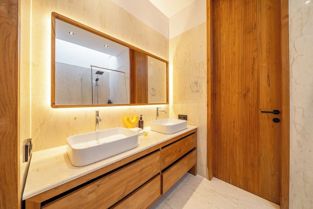 A modern bathroom with a large mirror, two sinks, wooden cabinetry, and a wooden door.